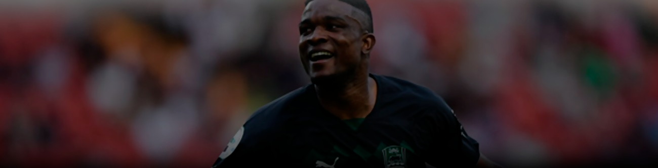 На изображении футболист в тёмной форме бежит по полю, вокруг него расплывчатый фон стадиона с размытыми зрителями. Он выглядит радостным, возможно, празднует забитый гол или успешное действие в игре