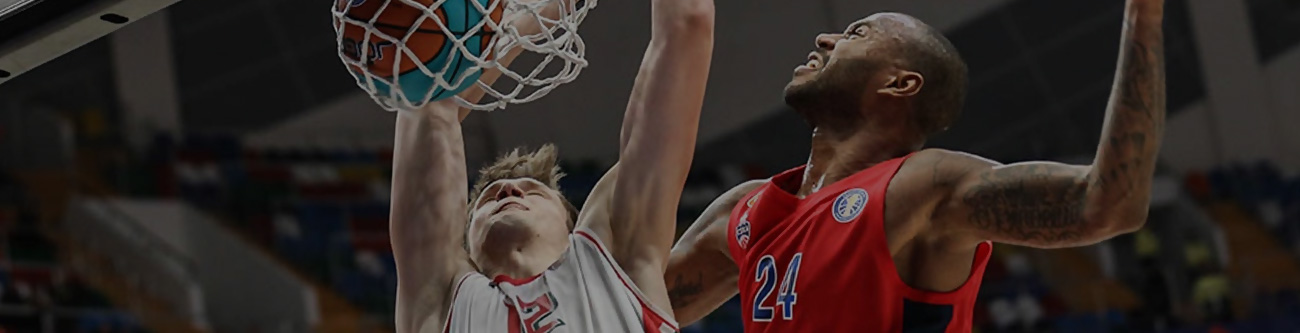 На изображении два баскетболиста в момент прыжка у корзины. Игрок слева в белой форме пытается забросить мяч, а игрок справа в красной форме пытается блокировать бросок. Видна напряжённая борьба под кольцом